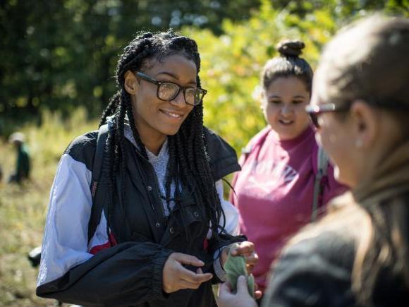 一位戴着黑框眼镜的女学生微笑着与她的同学交谈，同学们正在绘制入侵物种在田野中的生长情况