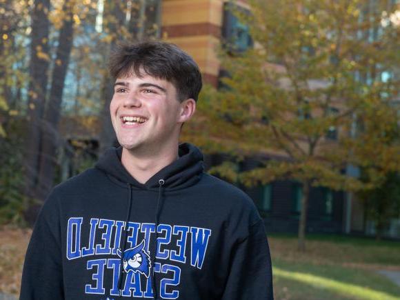 Student wearing black WSU sweatshirt with blue writing and owl logo smiling with fall trees in background.