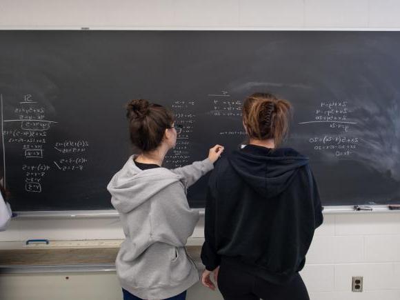 Two students with pony tails and hooded sweatshirts doing math equations on chalkboard
