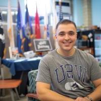 Ethan Lichwan, 2025届毕业生. 他在学校的退伍军人和军事办公室工作, 穿着一件灰色衬衫，胸前印有“华盛顿州立大学”的蓝色字样. 他的头发剪得很短. 背景中的彩旗模糊不清，他坐在一张绿色的沙发上.