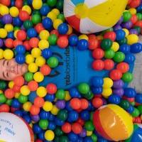 A male student from the 2023 Fresh Check Day. He is wearing a dark blue shirt with black lettering that says, "Fresh Check Day", and is laying in a colorful ball pit while smiling at the camera.